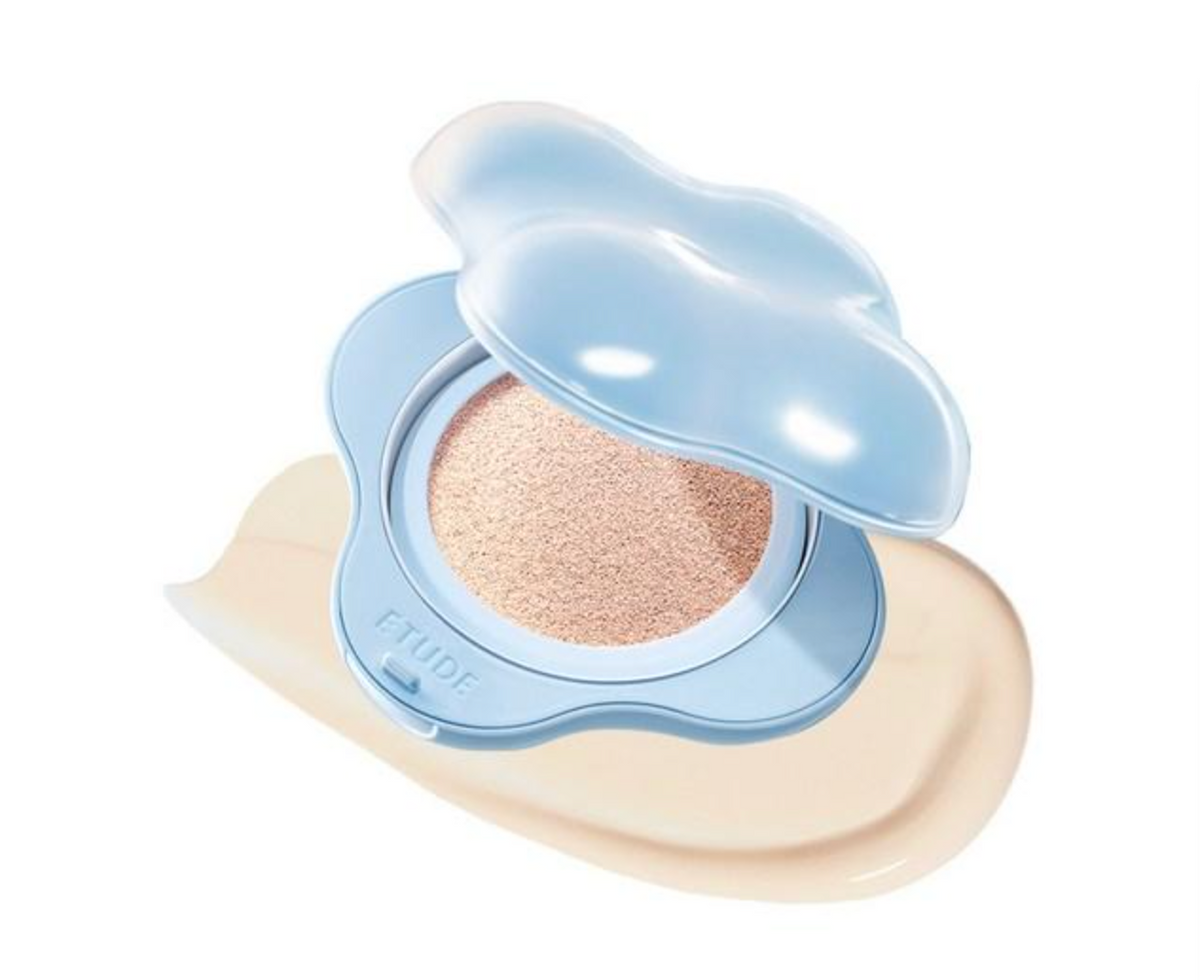 A cloud-shaped cushion foundation compact by CYUDE, shown open with the light blue lid propped up, revealing a beige-toned powder cushion. A smear of the beige foundation product is visible beneath the compact on a clean white background.