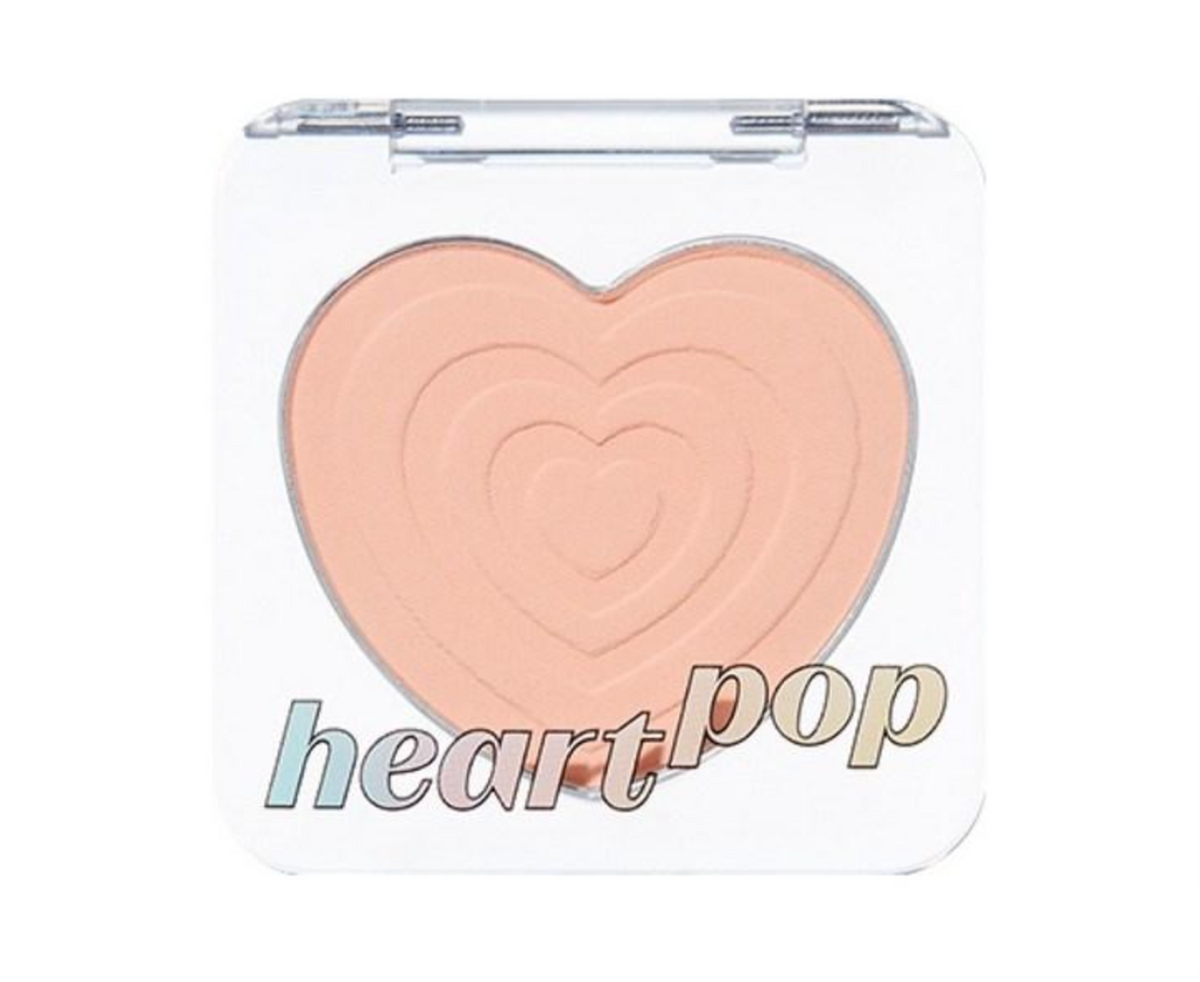 A makeup compact with a clear lid and a large heart-shaped pressed powder in a peachy beige shade. The words 'heart pop' are printed across the lower front of the compact.