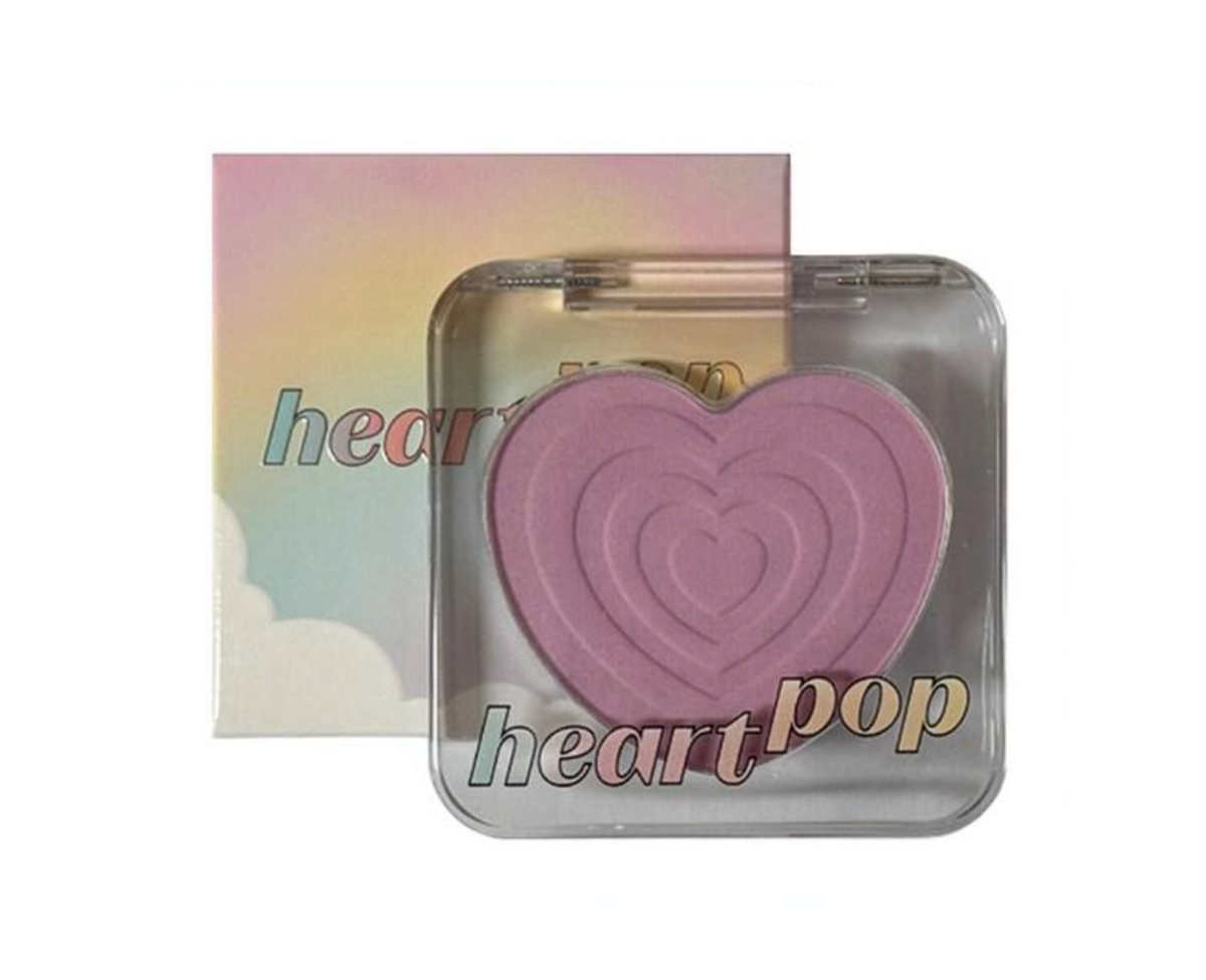 A heart-shaped pressed powder makeup compact in front of its pastel rainbow box. The compact has a clear lid and features the text 'heart pop' in a stylized font.