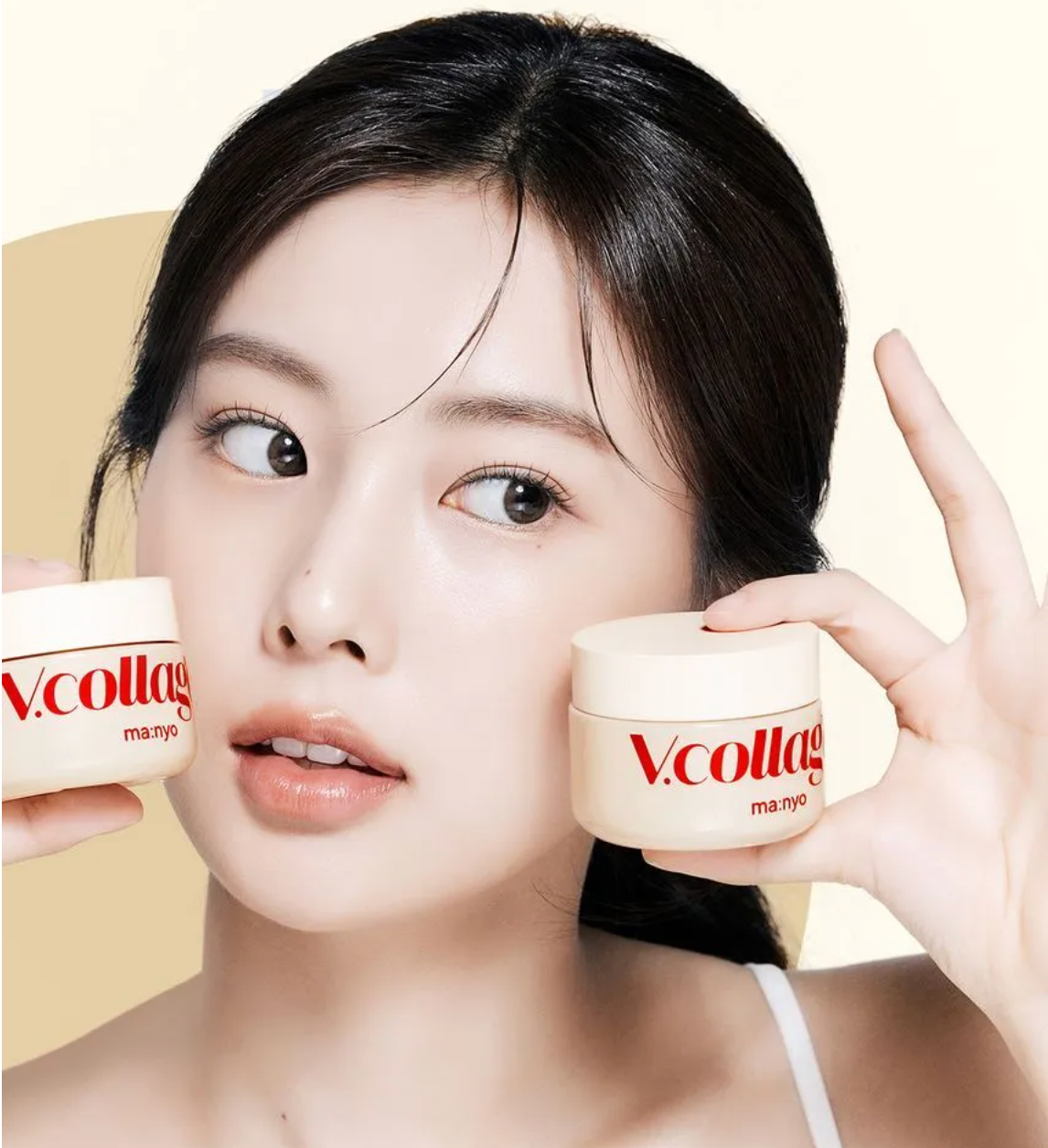 A close-up of a woman holding two cream-colored cosmetic jars branded 'V.collage' and 'ma:nyo' against a soft beige background.