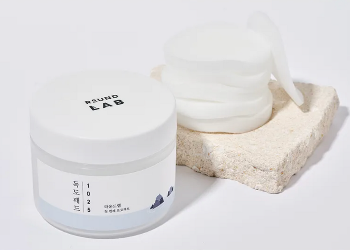 A white cosmetic jar labeled ROUND LAB 1025 with a stack of white sheet masks resting on a textured beige stone slab against a clean white background.