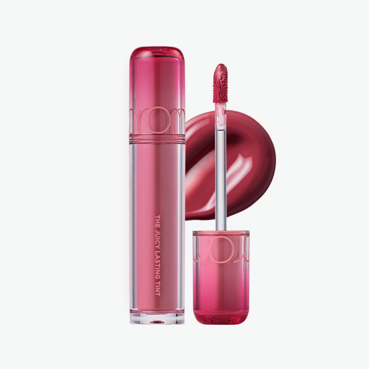 A clear tube of liquid lipstick with a translucent pink cap stands next to its cap and a doe-foot applicator loaded with a deep berry pink shade. A circular swatch of the lipstick color is shown in the background.