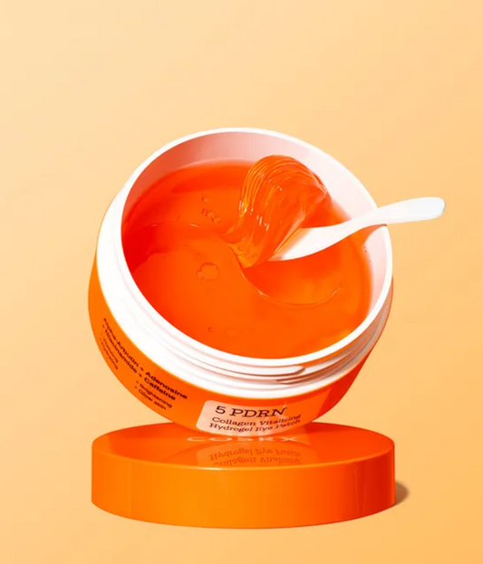 An open white cosmetic jar filled with bright orange gel, with a white spatula resting inside. The jar has an orange label and sits on a matching orange lid against a warm peach background.