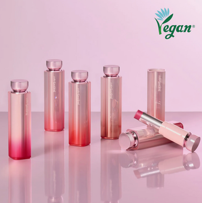 Six Vegan cosmetics bottles and tubes in a metallic pink-to-rose gradient finish, arranged on a reflective surface against a soft pink background. One tube is open, showing a pink product inside.