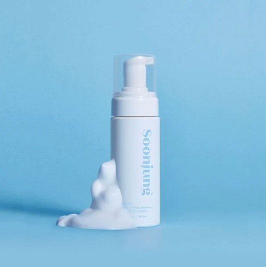 A white Soonjung foaming cleanser bottle with a clear cap stands on a light blue surface with white foam dripping from the base. The brand name 'soonjung' is printed vertically in blue on the bottle against a solid light blue background.