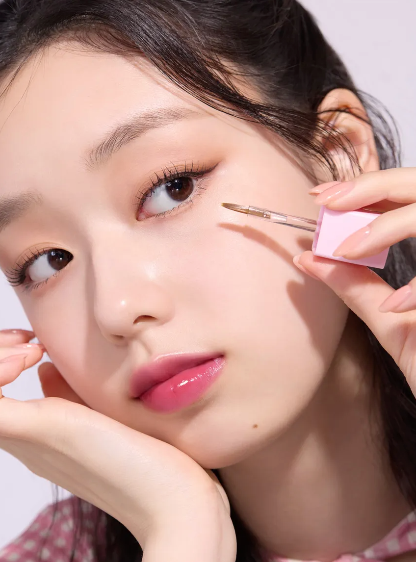 A close-up lifestyle shot of a woman applying liquid eyeliner with a pink-capped applicator wand. She has on a glossy pink lip and subtle eyeshadow, with her hand holding the applicator near her eye against a clean white background.