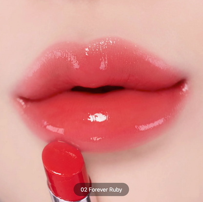 Close-up of glossy lips wearing a coral pink lip color, with a red lipstick bullet at the bottom and a label reading '02 Forever Ruby'.