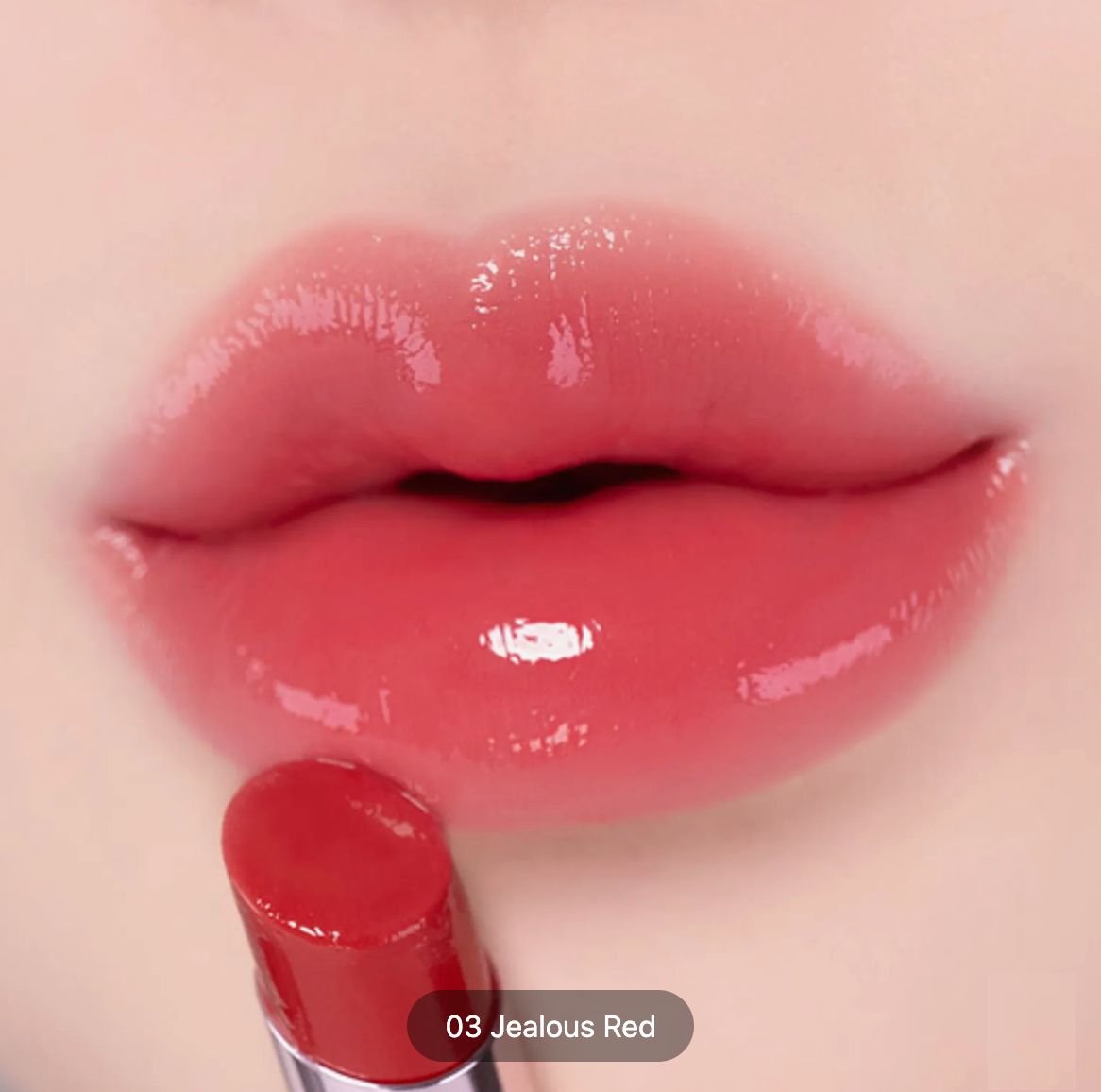 Close-up of glossy lips wearing a coral-red lip color, with a matching red lipstick bullet touching the lower lip. Text at the bottom reads '03 Jealous Red'.