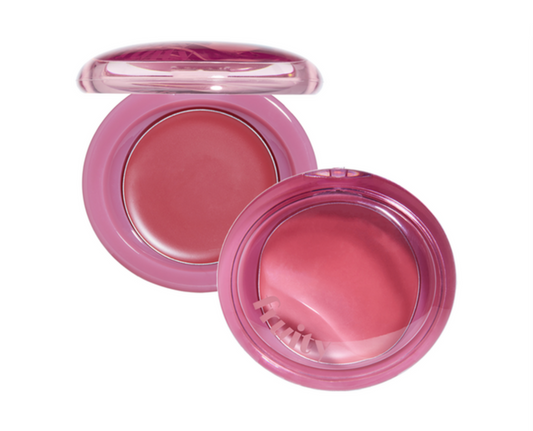 An open round pink makeup compact showing a rosy pink pressed powder, with the lid placed above and a small applicator brush resting inside, on a plain white background.