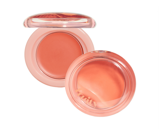An open round makeup compact showing a peachy coral pressed powder, with a translucent pink case and a small applicator brush visible inside, on a plain white background.