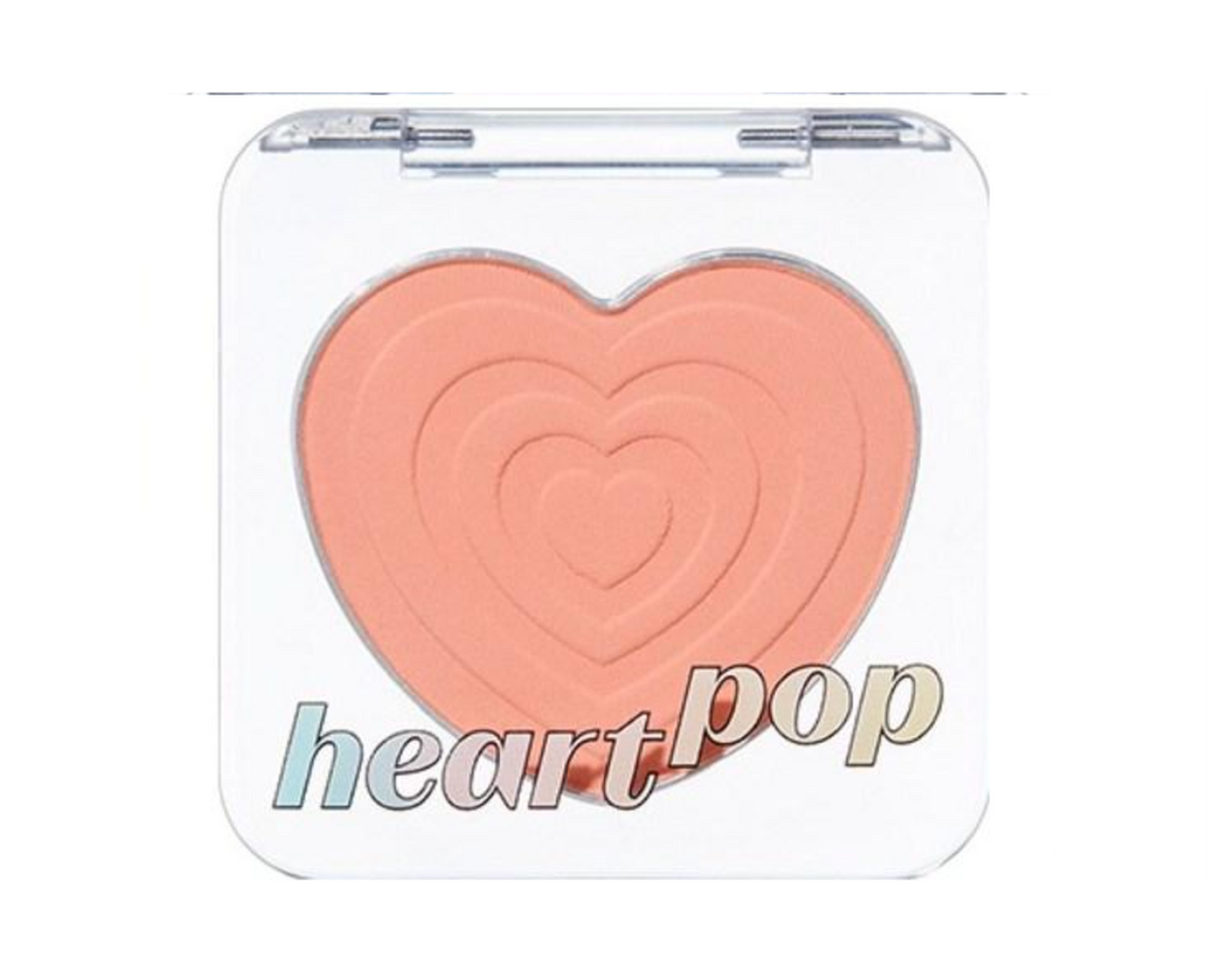 A makeup compact with a clear lid and a large heart-shaped pressed powder in a peachy pink shade. The words 'heart pop' are printed across the lower front of the compact.
