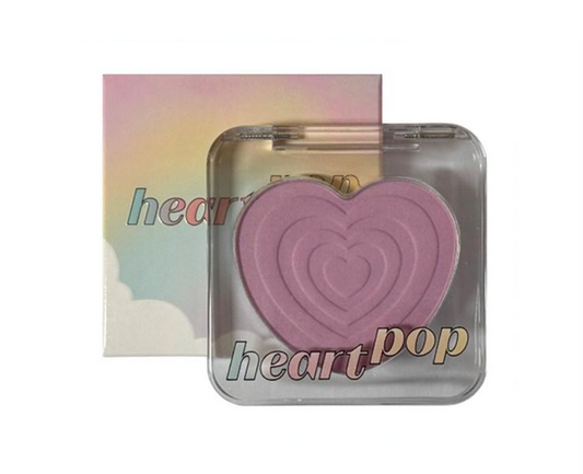 A heart-shaped pressed powder makeup compact in front of its pastel rainbow box. The compact has a clear lid and features the text 'heart pop' in a stylized font.
