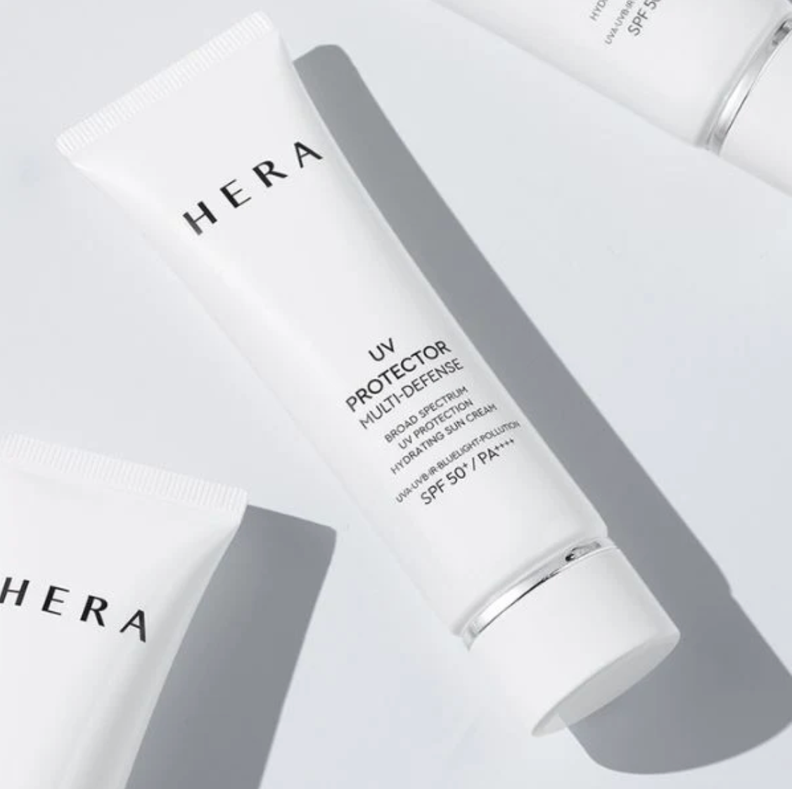 White HERA UV Protector Multi-Defense SPF 50+ sunscreen cream tube, shown in a clean, modern flat-lay composition with soft gray shadows on a light gray background.