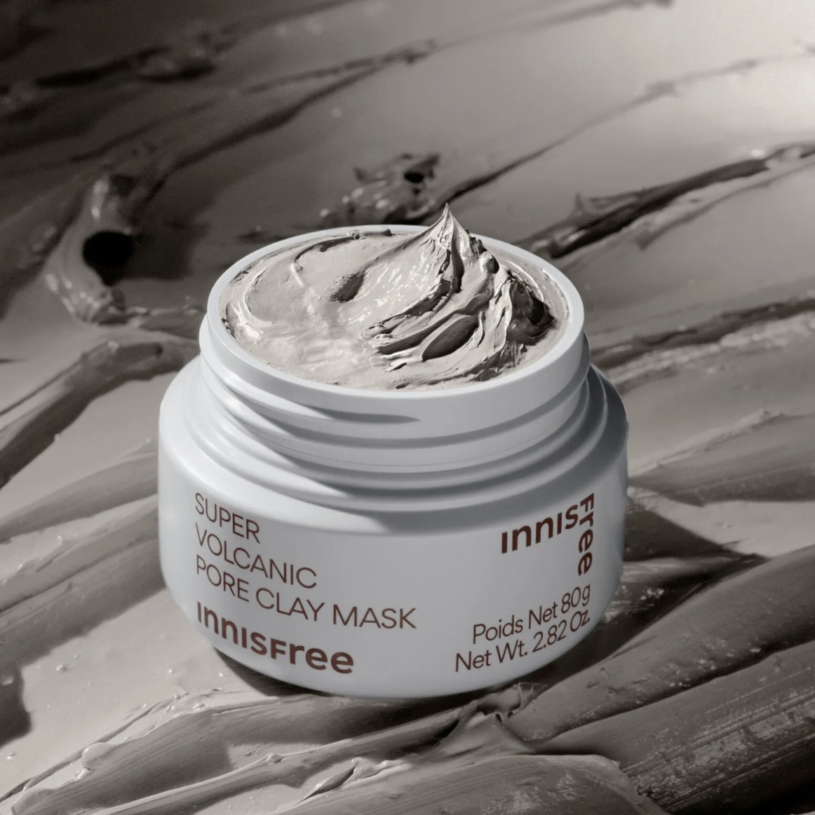 An open white jar of Innisfree Super Volcanic Pore Clay Mask, showing the thick gray clay mask inside, set against a textured background of the same gray mask cream.