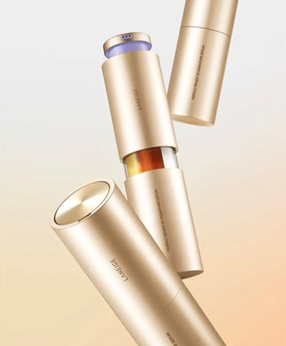 Three metallic gold cosmetic tubes floating diagonally on a soft beige-to-white gradient background. One tube is open, revealing a small amber bottle inside, and the brand name LANEIGE is printed vertically on the tubes.
