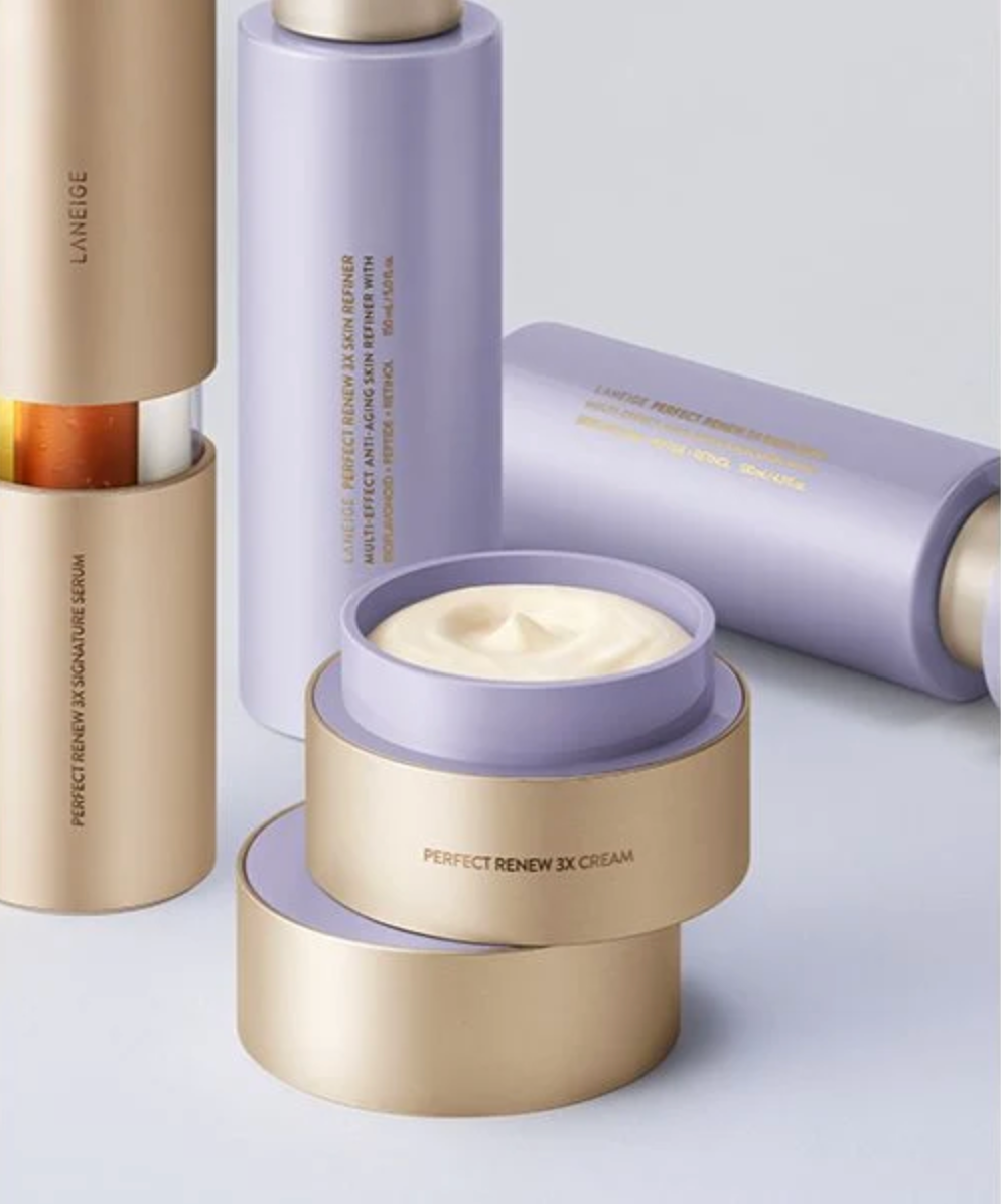 A close-up studio shot of several metallic rose-gold cosmetic cream jars stacked together, with a thick white cream swatch placed in front of the center jar. The visible text on the jars reads 'PERFECT RENEW 3X CREAM'.