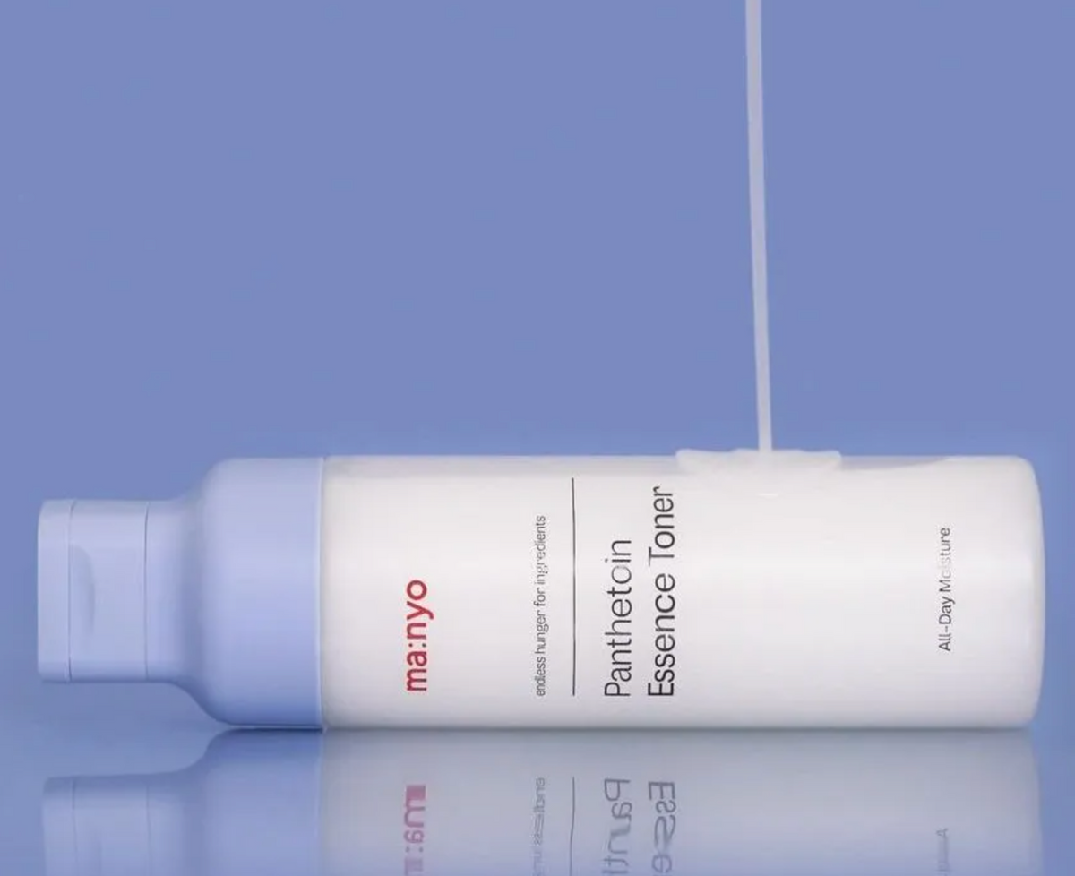 A white bottle of ma:nyo Panthetoin Essence Toner with a light blue cap, shown horizontally on a reflective surface against a solid periwinkle blue background. A single drop of clear serum is dripping from the top of the bottle.