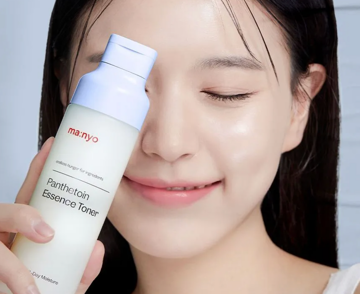 A woman with closed eyes holds a bottle of manyo Panthetoin Essence Toner near her face. The bottle is white with a light blue cap, and the background is a soft, out-of-focus light blue.