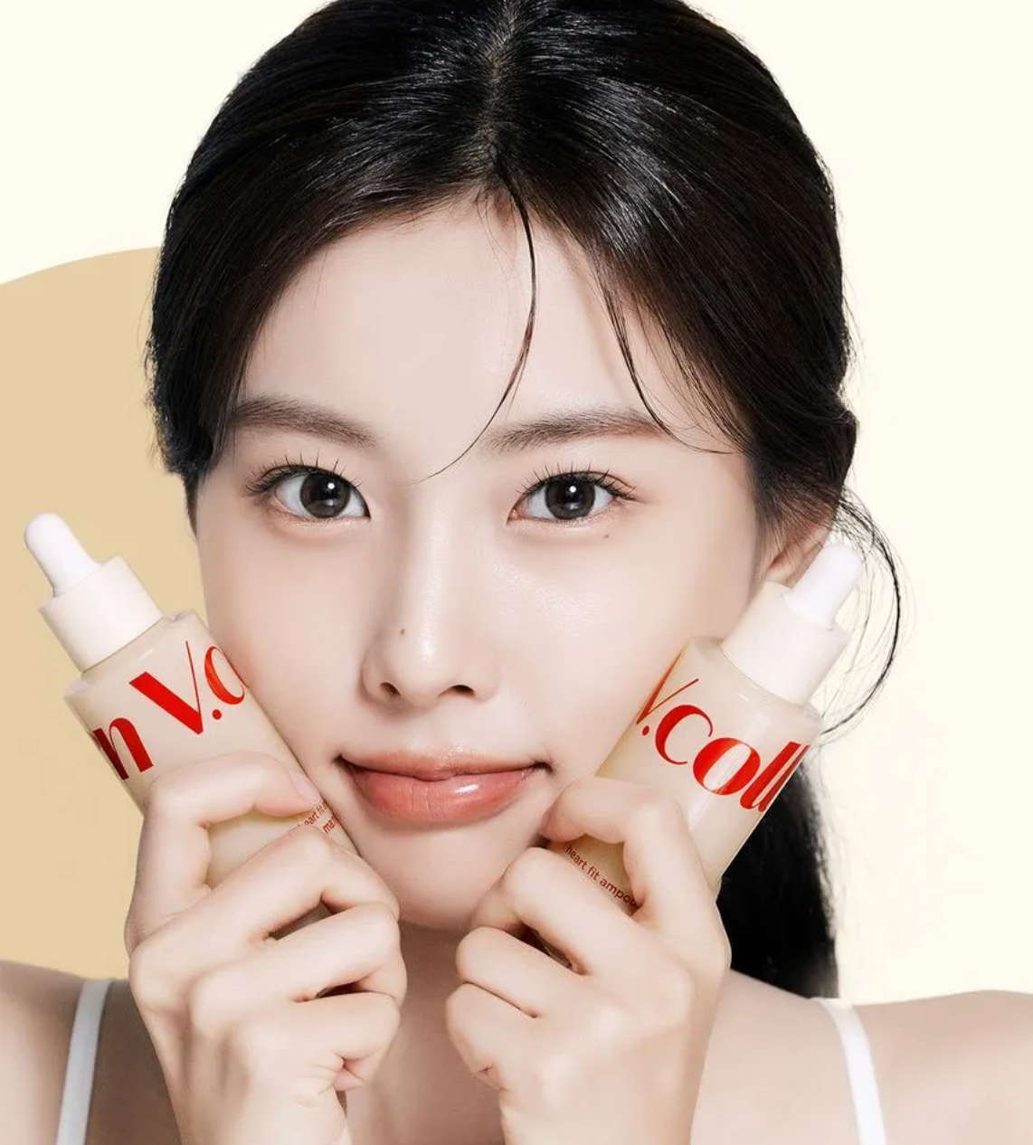 A close-up lifestyle shot of a woman holding two cosmetic dropper bottles near her cheeks. The bottles are frosted with a warm beige tint and feature red branding text.