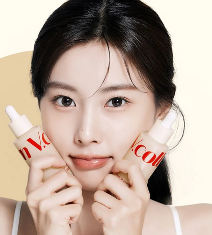 A close-up lifestyle shot of a woman holding two cosmetic dropper bottles near her cheeks. The bottles are frosted with a warm beige tint and feature red branding text.