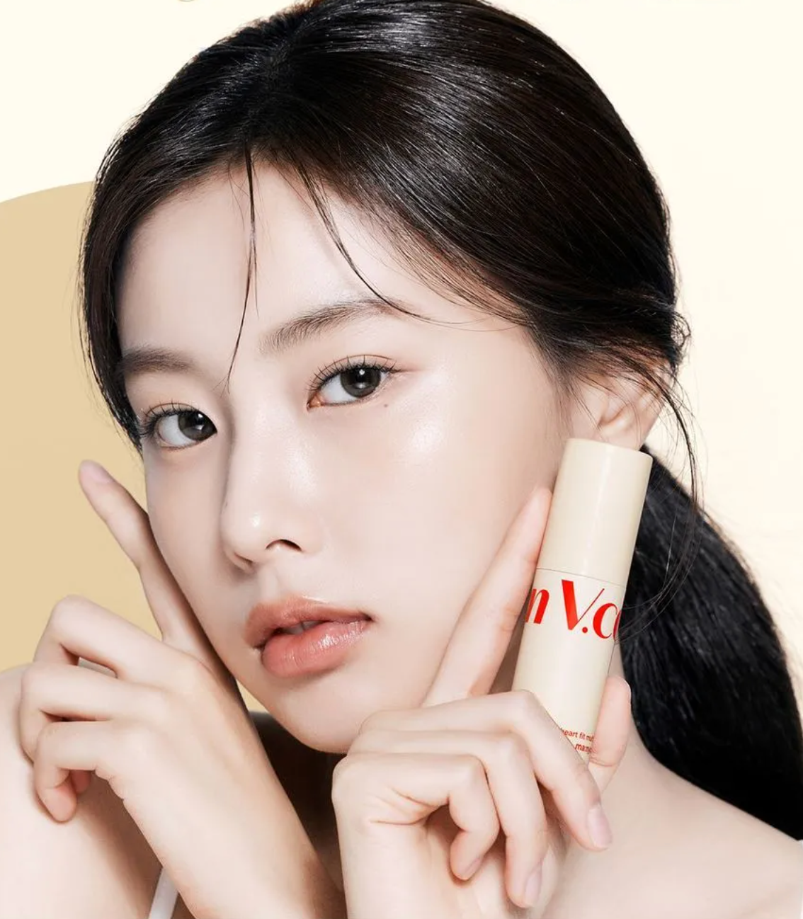 A close-up beauty shot of a woman holding a beige cosmetic stick container with red lettering against a soft beige background.