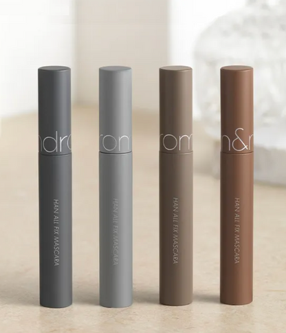 Four Han All Fix Mascara tubes in different neutral shades (gray, taupe, brown, and medium brown) lined up on a light beige surface. 
