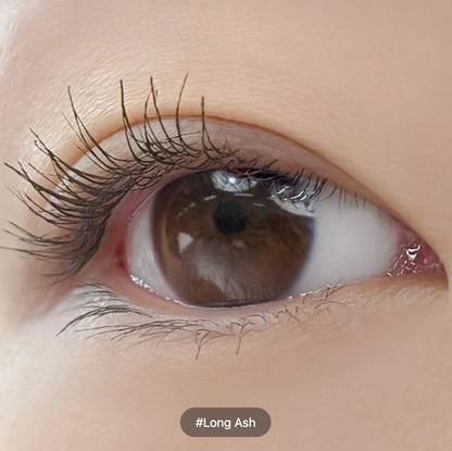 Extreme close-up of a human eye with long, dark ash brown eyelashes and a brown iris, with a skin-toned background and a label reading '#Long Ash' at the bottom.
