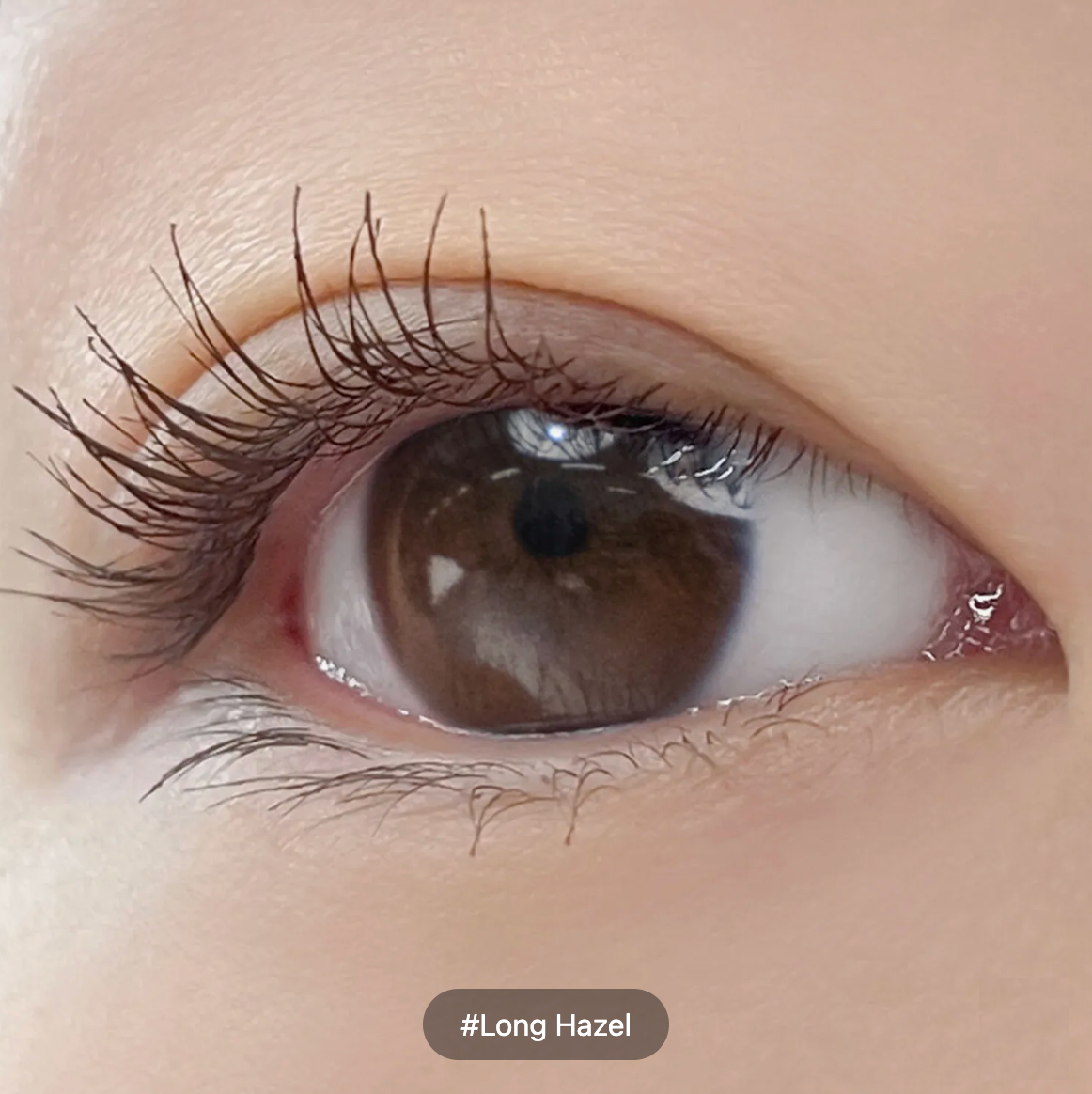 Extreme close-up of a long, dark brown hazel eye with long eyelashes and a soft peach-toned eye area. A small label at the bottom reads '#Long Hazel'.