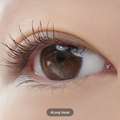 Extreme close-up of a long, dark brown hazel eye with long eyelashes and a soft peach-toned eye area. A small label at the bottom reads '#Long Hazel'.