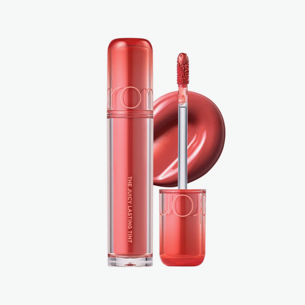 A tube of UOM lip gloss in a translucent coral-red shade, shown with the cap off, the applicator wand, and a circular swatch of the gloss on a clean white background.