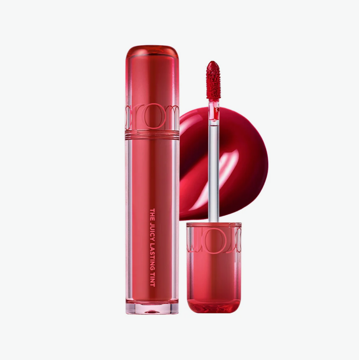 A red liquid lipstick tube and its applicator wand, shown on a clean white background with a glossy red lipstick smear behind them. The brand name 'rom' is embossed on the tube.