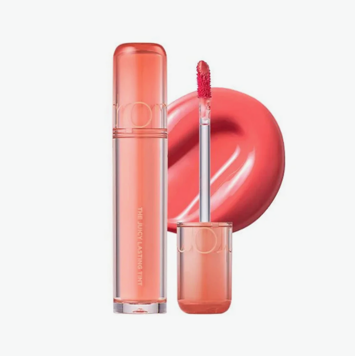 A clear cosmetic tube containing a peachy coral liquid lip product, shown with the cap removed and a doe-foot applicator. A large circular swatch of the same coral-red shade appears behind the product on a clean white background.