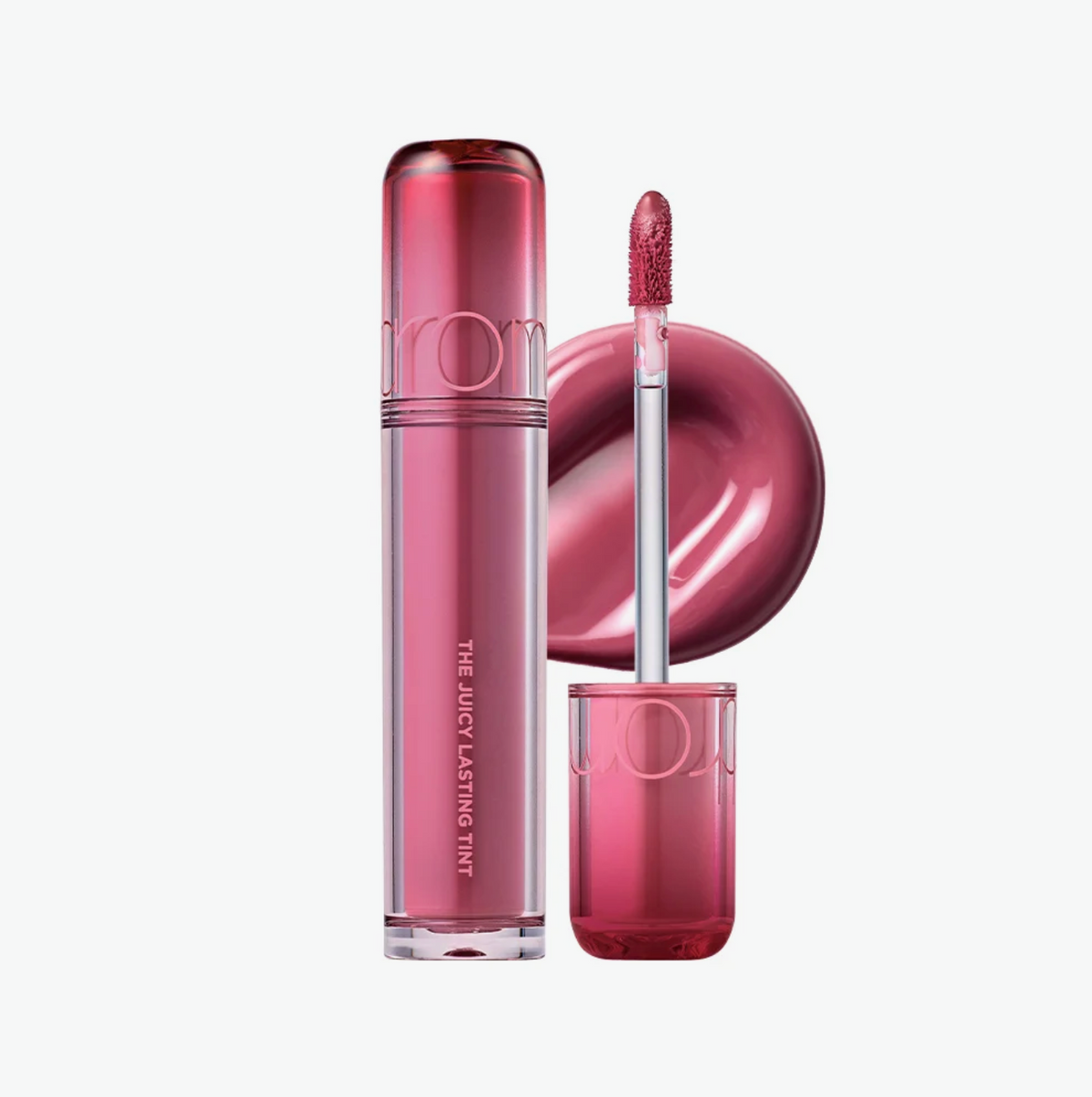 A tube of lip tint next to its applicator wand and a circular color swatch, all in a glossy metallic pink packaging on a light gray background.