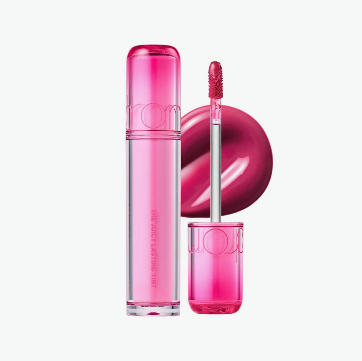 A pink lip gloss tube and its applicator wand are displayed on a clean white background, with a glossy circular swatch of the pink gloss shown behind them. The brand name 'rom' is embossed on the tube and cap.