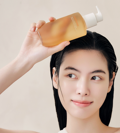 A woman with wet, straight black hair holds a translucent amber pump bottle above her head against a soft beige background.