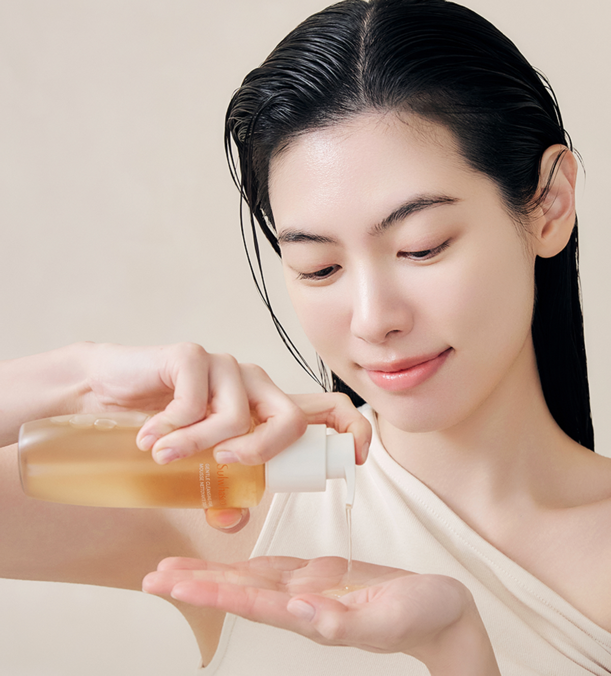 A woman with wet, styled black hair applies a clear pump bottle containing a golden liquid to her palm in a clean, close-up beauty shot against a soft beige background.