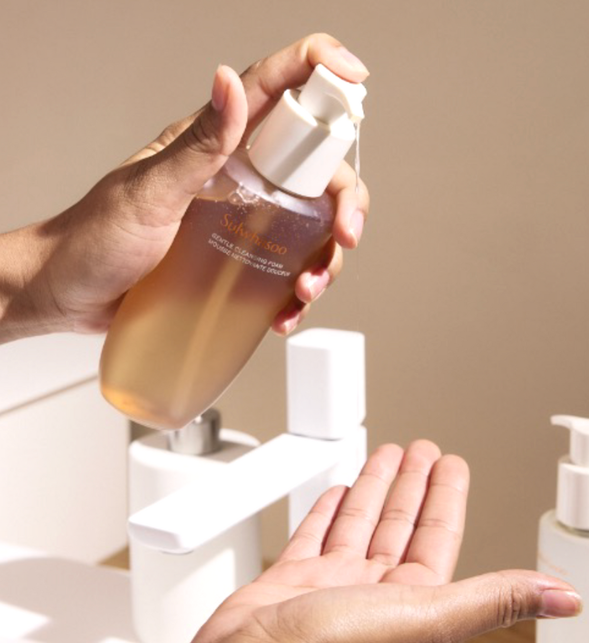 A hand holding a frosted bottle filled with amber liquid and dispensing product onto another hand, with a white faucet and pump bottle in the background.