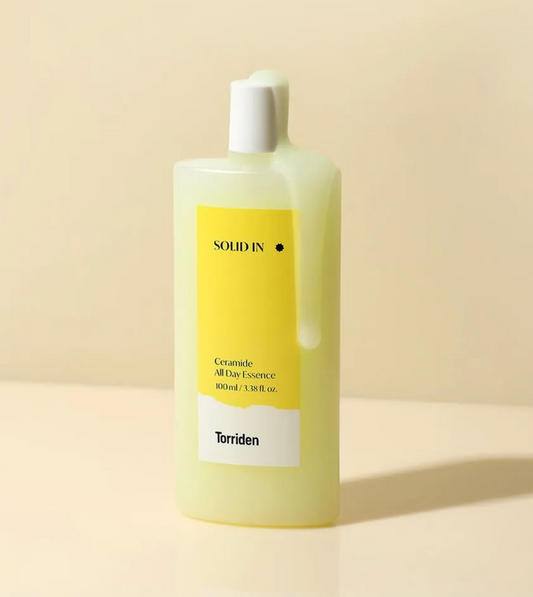 A bottle of Solid In Ceramide All Day Essence by Torriden, featuring a pale yellow translucent bottle with a white cap and a yellow and white label, set against a warm beige background.